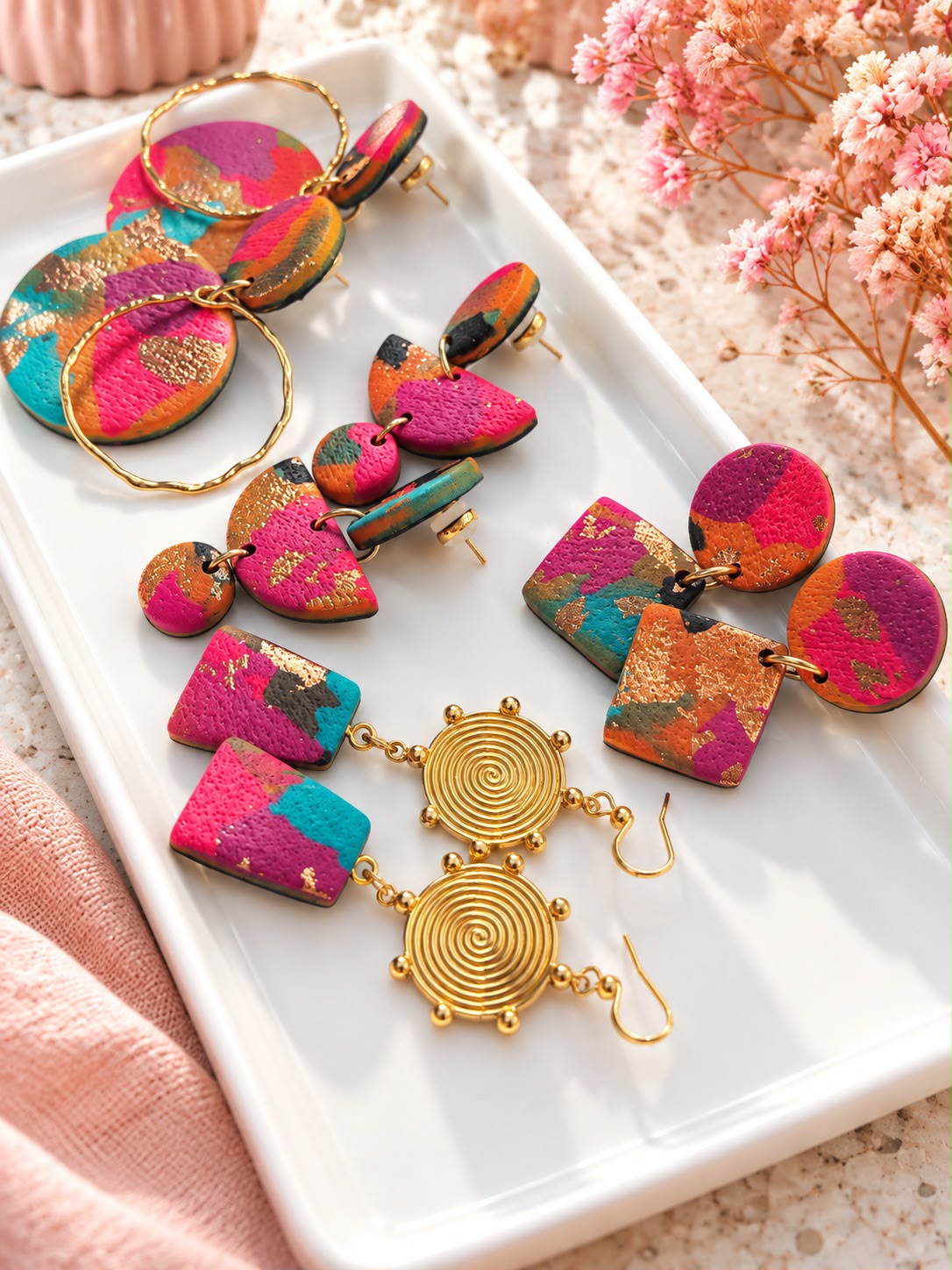 Quatre paires de boucles d'oreilles pendantes en argile polymère fuchsia, turquoise et orange à la feuille dorée, sur un plateau blanc