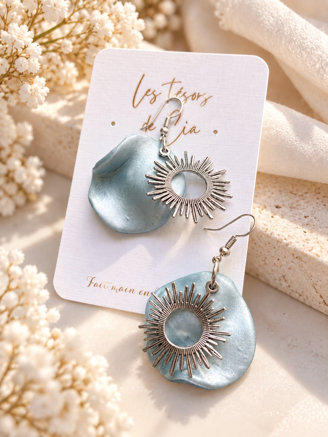 Boucles d'oreilles à crochet, pétale en argile polymère bleu nacré et soleil rayonnant argenté, sur une carte Les Trésors de Cia
