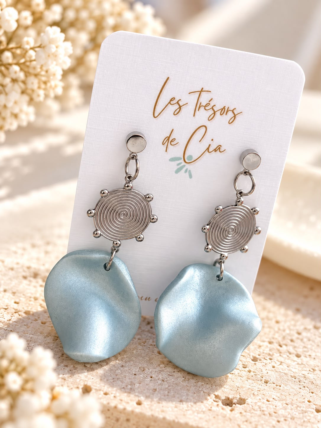 Boucles d'oreilles pendantes en argile polymère bleu ciel nacré, disque spirale argenté et puce ronde, posées sur une carte Les Trésors de Cia