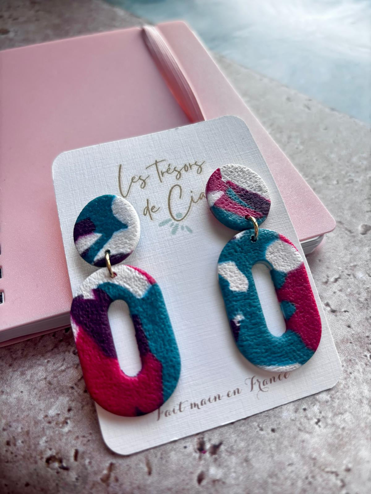 Boucles d'oreilles ovales ajourées en argile polymère traversée de larges taches bleu canard, rose fuchsia, violet prune et blanc, avec une puce ronde en haut et une fente allongée au centre du pendant.