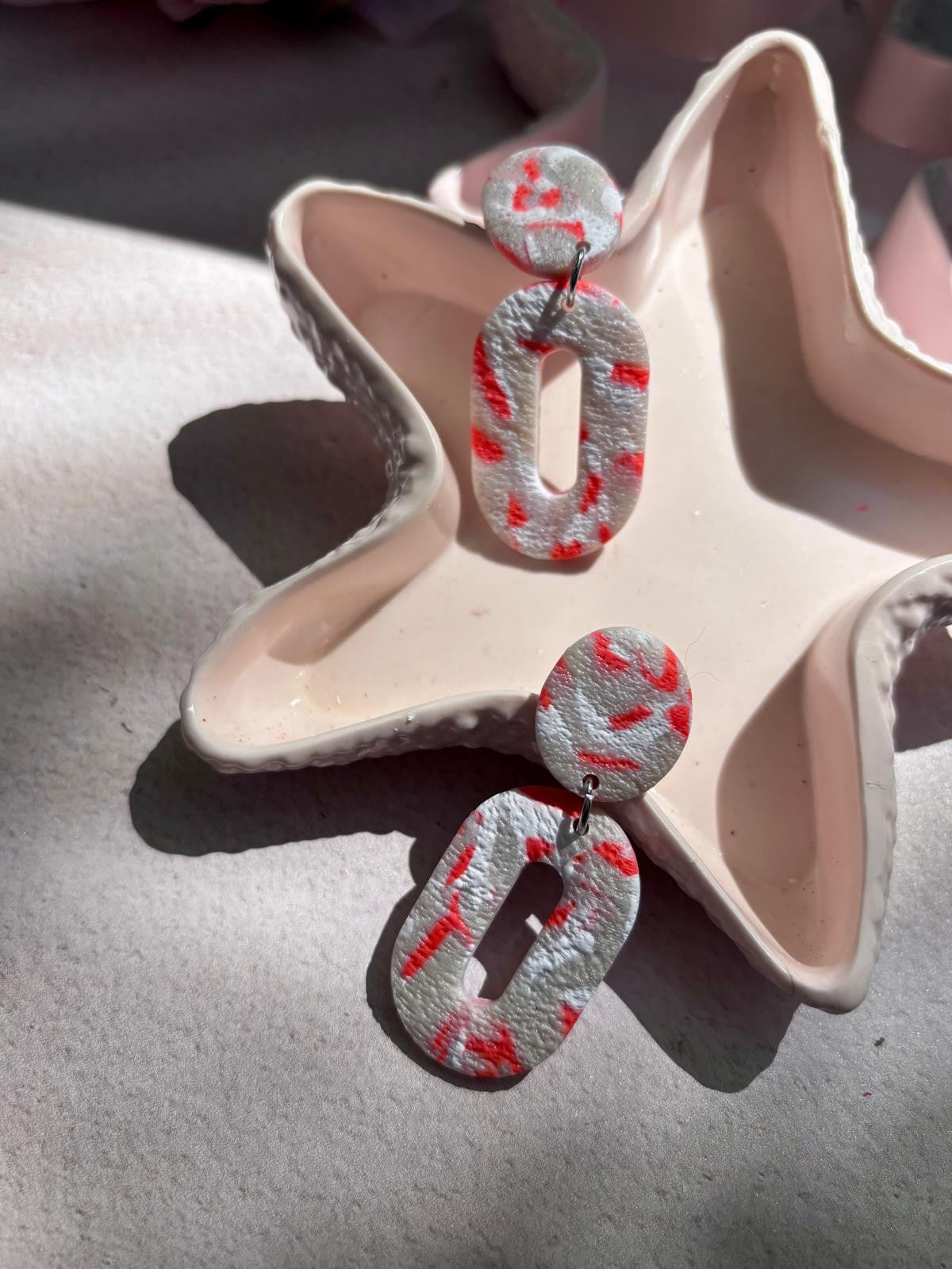 Boucles d'oreilles ovales ajourées en argile blanche texturée parcourue de mouchetures rouge corail, puce ronde et fente allongée au centre du pendant ovale.