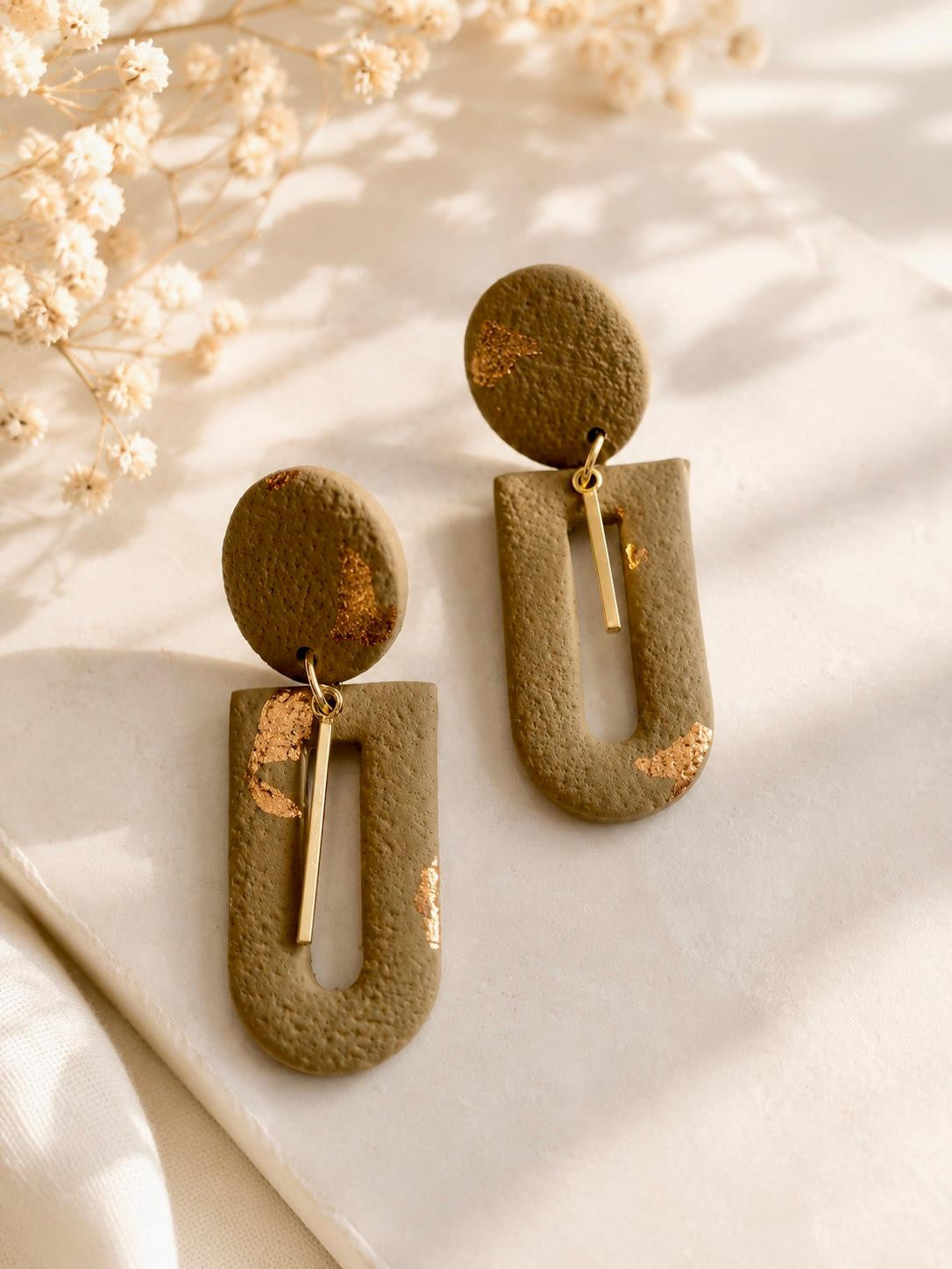 Boucle d'oreille en arche évidée en argile vert kaki mat aux éclats cuivrés, avec une fine barre dorée suspendue dans l'ouverture