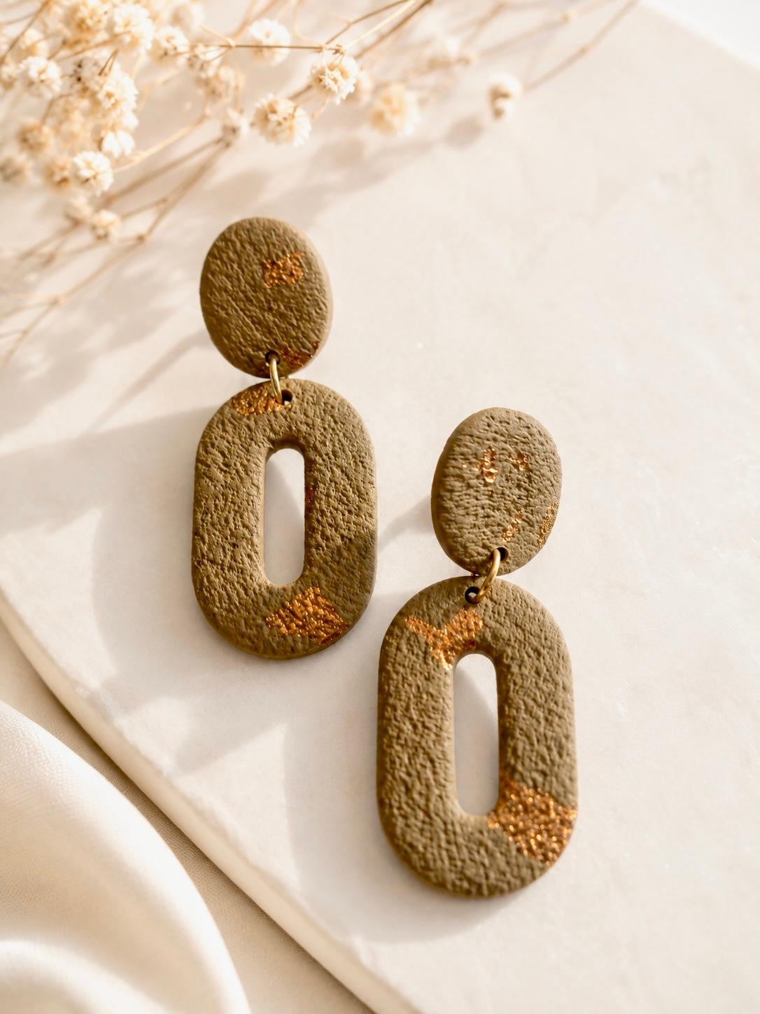 Boucles d'oreilles ovales ajourées en argile vert kaki mat, avec des éclats de feuille cuivrée par endroits et une fente allongée au centre du pendant.