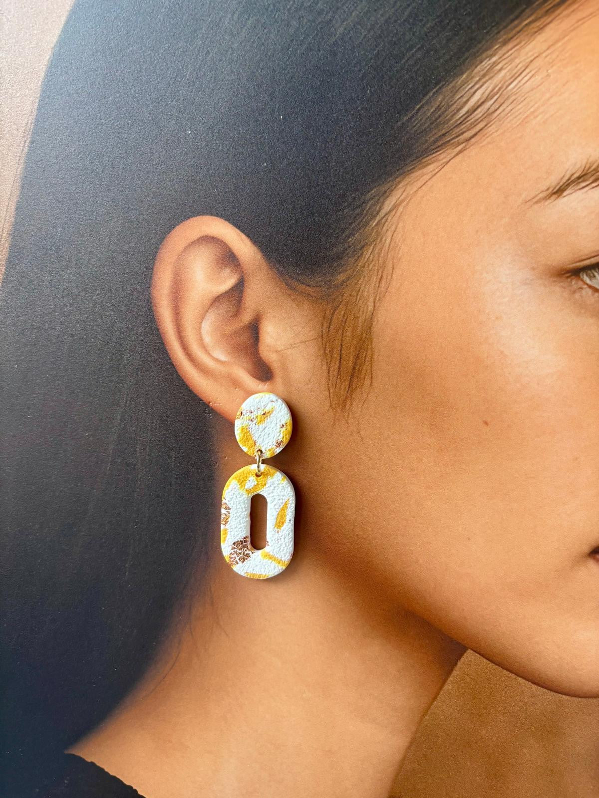 Boucles d'oreilles ovales ajourées en argile blanche traversée de coulées jaune vif, avec une fente allongée au centre du pendant.