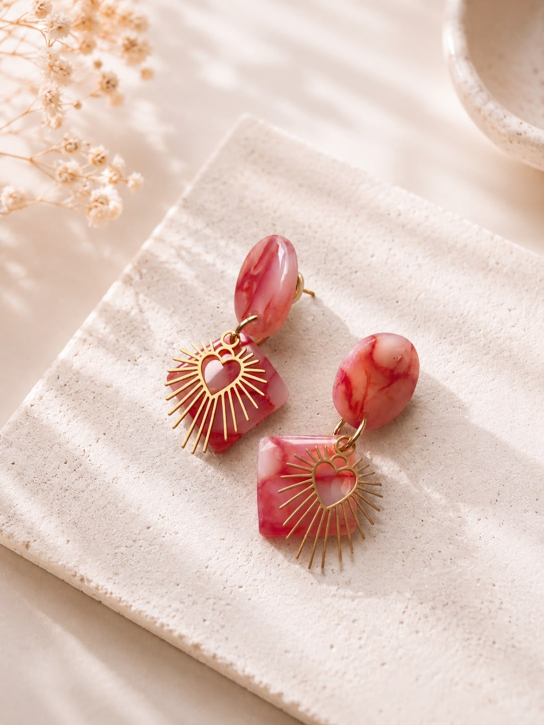 Paire de boucles d'oreilles carré marbré rose et cœur doré rayonnant, posée sur une pierre claire